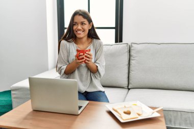 Genç Latin kadın kahvaltısını yapıyor. Evdeki kanepede oturuyor.