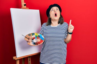 Orta yaşlı İspanyol kadın paletle resim çiziyor. Şaşkın şaşkın duruyor ve parmaklarıyla işaret ediyor ve kollarını kaldırıyor.. 