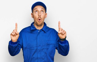 Sakallı, inşaatçı tulumu giyen kel adam şaşırmış ve şaşırmış bir şekilde yukarı bakıyor ve parmaklarıyla işaret ediyor ve kollarını kaldırıyor.. 