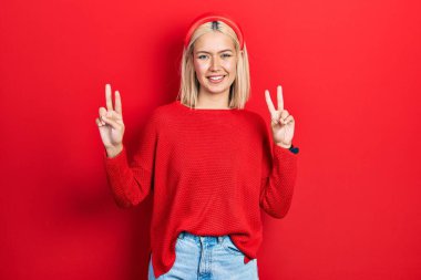 Gayri resmi kırmızı süveter giyen güzel sarışın kadın kameraya bakıp zafer işareti yapıyor. İki numara.. 