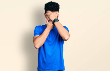 Sakallı, günlük mavi tişörtlü, gözleri ve ağzı elleri kapalı, şaşırmış ve şok olmuş genç bir Arap. duygu gizleme 