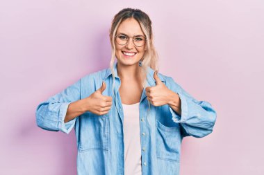 Güzel sarışın kadın, günlük kıyafetler ve gözlük takıyor. Başarı işareti yapıyor. Elleriyle olumlu jest yapıyor. Neşeli ifade ve kazanan jest. 