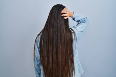 Genç İspanyol kadın mavi arka planda geriye doğru duruyor. Elleri kafasında şüpheyi düşünüyor. 