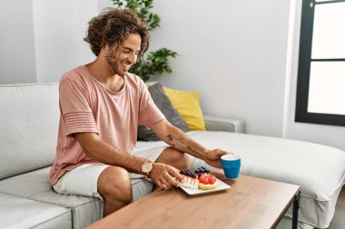 Genç İspanyol adam kahvaltısını yapıyor. Evdeki kanepede oturuyor..