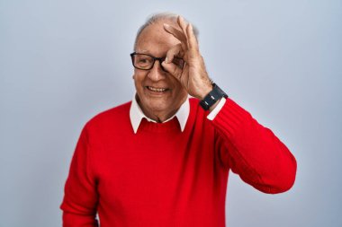 Gri saçlı yaşlı adam izole edilmiş arka planda duruyor. El hareketleriyle gülümsüyor, gözleriyle mutlu yüzüyle parmaklarının arasından bakıyor.. 