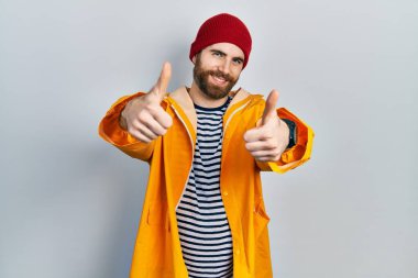 Sakallı, sarı yağmurluk giyen, elinde olumlu bir jest yapan, gülümseyen ve başarı için sevinen beyaz bir adam. kazanan jest. 