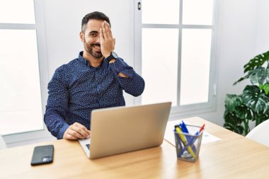 Ofiste sakallı, laptopla çalışan, bir gözünü elinle kapatan, yüzünde kendine güvenen bir gülümseme ve sürpriz duygular olan genç İspanyol bir adam.. 