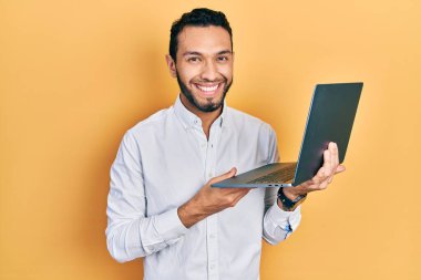 Sakallı İspanyol bir adam bilgisayarla çalışıyor. Pozitif ve mutlu görünüyor. Kendine güvenen bir gülümsemeyle dişlerini gösteriyor. 