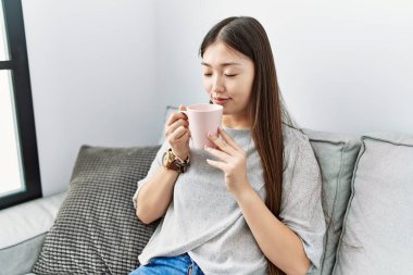 Genç Asyalı kadın oturma odasındaki kanepede kahve içiyor.