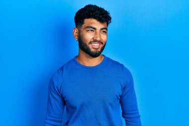 Sakallı, sıradan mavi süveter giyen, yüzünde gülümseme, doğal bir ifade olan Arap bir adam. Kendinden emin gülen. 