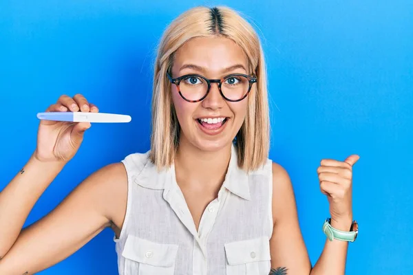 Beautiful blonde woman holding pregnancy test result pointing thumb up to the side smiling happy with open mouth