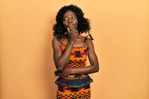 若いアフリカ系アメリカ人の女性は 伝統的なアフリカの服を着て 顎の質問 集中的な表現について考えています 思慮深い顔で笑顔 疑わしい概念 — ストック写真