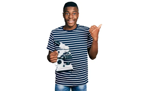 Young African American Man Holding Microscope Pointing Thumb Side Smiling — Stock Photo, Image