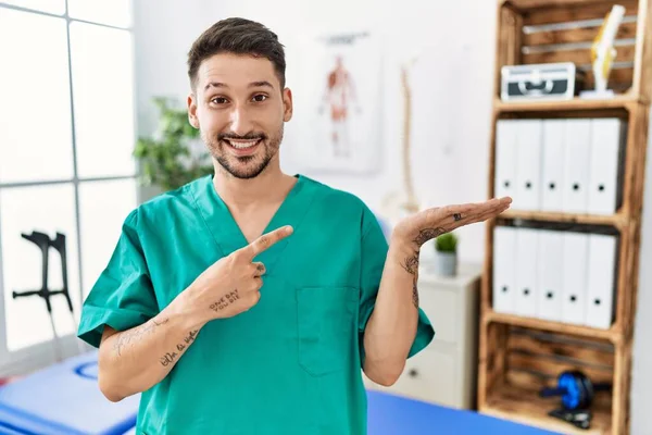 若い理学療法士の男は手で提示しながら カメラに驚いて笑顔と指で指して回復クリニックで働いています — ストック写真