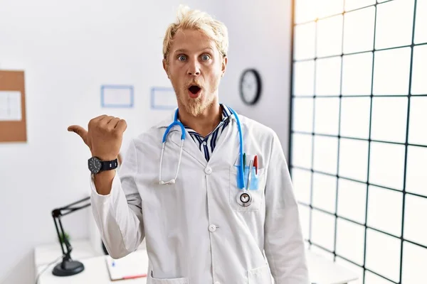 Jonge Blonde Man Doktersuniform Stethoscoop Kliniek Verrast Wijzend Met Hand — Stockfoto