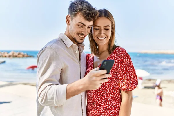 若いですヒスパニックカップル上の休暇笑顔幸せな使用してスマートフォンでビーチ — ストック写真