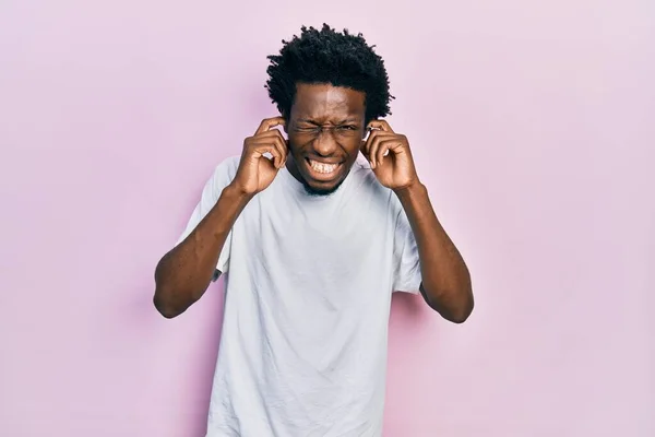 カジュアルな白いTシャツを着た若いアフリカ系アメリカ人男性は 大きな音楽のノイズに悩まされた表情で耳を覆います 聴覚障害の概念 — ストック写真