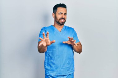 Sakallı, mavi erkek hemşire üniforması giyen yakışıklı, tiksindirici ve tiksindirici bir yüz ifadesi takınan, tiksinti reaksiyonu yüzünden tiksinmiş bir İspanyol. Eller havada. 