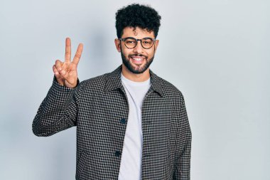 Sakallı, gözlüklü genç bir Arap. İki numaralı parmaklarıyla işaret ediyor. Kendinden emin ve mutlu bir şekilde gülümsüyor.. 