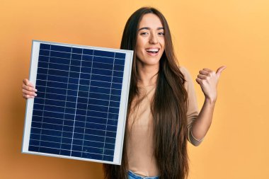 Fotovoltaik güneş paneli tutan genç İspanyol kız başparmağını yana uzatıyor ve ağzı açık gülümsüyor. 