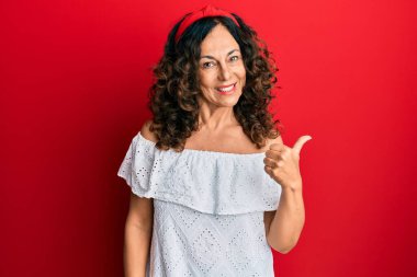 Orta yaşlı İspanyol kadın, günlük kıyafetler giyiyor, gülümsüyor, gülümsüyor ve başparmağıyla yan tarafa bakıyor.. 