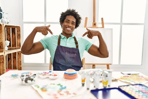 年轻的非洲裔美国人坐在艺术工作室的桌子上 面带微笑 满脸自信 用手指指着自己 自豪而快乐 — 图库照片