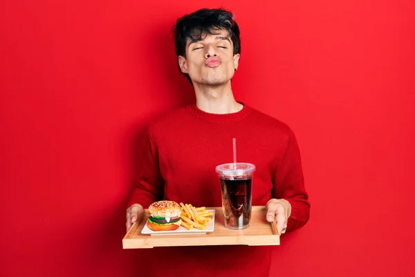 Handsome hipster young man eating a tasty classic burger with fries and soda looking at the camera blowing a kiss being lovely and sexy. love expression.