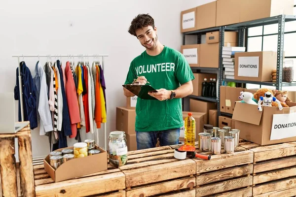 Young Hispanic Man Wearing Volunteer Uniform Writing Clipboard Charity Center — Stockfoto