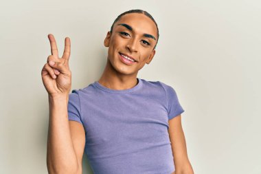 Makyaj yapan, uzun saçlı, sıradan tişörtlü, iki numaralı parmaklarıyla gösteriş yapan, kendinden emin ve mutlu gülümseyen İspanyol bir adam.. 