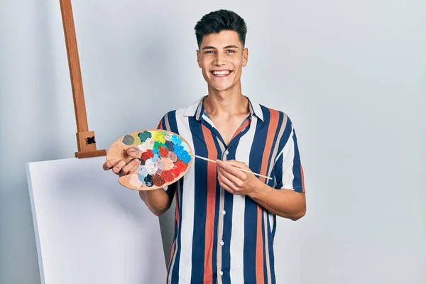 画家のパレットと筆を持っている若いヒスパニック系の男は 顔に幸せとクールな笑顔で笑みを浮かべてキャンバスに近い立っている 歯を見せて — ストック写真