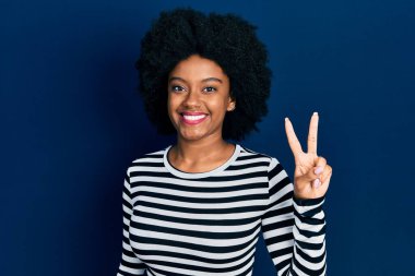 Genç Afro-Amerikalı kadın günlük kıyafetler giyiyor ve iki numarayı gösterip işaret ederken kendinden emin ve mutlu bir şekilde gülümsüyor.. 