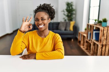 Genç Afro-Amerikan kadın, gündelik giysiler içinde evde oturmuş gülümsüyor ve el ve parmaklarıyla 