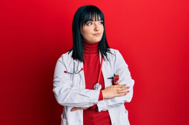 Doktor üniforması ve steteskop giymiş genç İspanyol kadın kollarını kavuşturup kendinden emin bir şekilde yan tarafa bakıyor. 