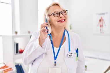 Doktor üniforması giyen orta yaşlı sarışın kadın klinikte akıllı telefondan konuşuyor.