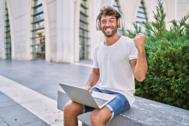 Kulaklık ve dizüstü bilgisayar kullanan genç İspanyol adam gururla bağırıyor, zaferi kutluyor ve başarıyı kollarını kaldırarak kutluyor. 