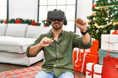 Genç İspanyol adam VR gözlük takıyor. Evdeki Noel ağacının yanında oturuyor.