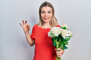 Güzel beyaz bir kadın elinde bir buket beyaz çiçek tutuyor tamam işareti yapıyor parmaklarıyla, gülümseyerek mükemmel bir sembol oluşturuyor. 