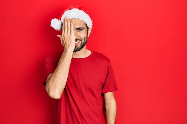 Mladý Hispánec Vánoční Čepici Zakrývající Jedno Oko Rukou Sebevědomý Úsměv — Stock fotografie
