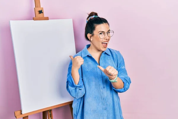 画家のイーゼルのそばに立つ若いヒスパニック系の女性は 手と親指を上げて後ろを指して立ち 自信を持って笑って — ストック写真