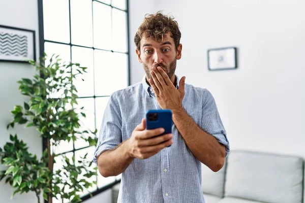 Νεαρός Όμορφος Άντρας Που Χρησιμοποιεί Smartphone Στο Σπίτι Καλύπτοντας Στόμα — Φωτογραφία Αρχείου