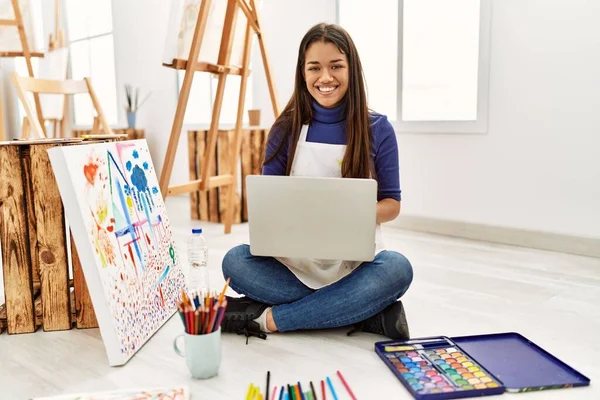 年轻的黑发女子坐在艺术工作室的地板上 膝上型电脑显得积极而快乐 面带微笑 露出自信的牙齿 — 图库照片