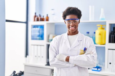 Afrika kökenli Amerikalı kadın bilim adamı üniforması giyiyor. Laboratuvarda kollarını kavuşturup el kol hareketi yapıyor.