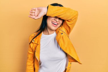 Güzel İspanyol kadın, burun piercingli, sarı deri ceketli, gözlerini kollarıyla kapatıp gülümseyen neşeli ve komik. kör kavram. 