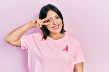 Tişörtüne pembe kanser kurdelesi takan genç İspanyol kadın barış sembolü yapıyor. Yüzünde parmaklar, gülümseyerek zaferi gösteriyor. 