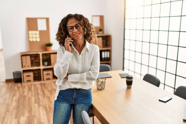 Orta yaşlı İspanyol kadın gülümsüyor ofisteki akıllı telefondan konuşuyor.