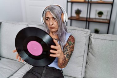 Orta yaşlı, gri saçlı, müzik dinleyen, elinde vinil plakla evde kuşkucu ve sinirli bir kadın, sorun yüzünden suratını asıyor. negatif kişi. 
