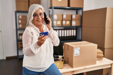 Orta yaşlı kadın ecommerce iş işçisi. Ofiste kredi kartıyla akıllı telefondan konuşuyor.