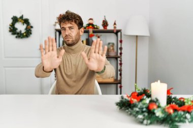 Masada sakallı genç yakışıklı bir adam Noel süslemesi ile oturuyor. Avuçlarını kaldırıyor, reddediyor ve iğrenç bir ifadeyle reddediyor. Dur ve yasakla. 