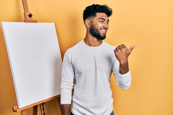 髭を生やしている男は画家によって立っているイーゼルスタンド親指を指している側に笑みを浮かべて幸せなオープン口 — ストック写真