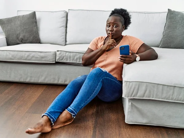 年轻的非洲女人坐在家里的地板上 用智能手机要求安静 手指放在嘴唇上 沉默和秘密概念 — 图库照片